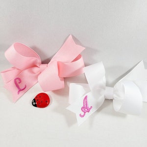 May include: Two hair bows, one pink and one white, with embroidered letters "L" and "A" on them. A red ladybug button is next to the pink bow.