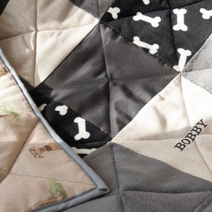 Puede incluir: Una colcha personalizada con un diseño de patchwork que presenta un estampado de huesos gris, negro y blanco, una tela beige con un estampado de perro y una tela beige con el nombre "BOBBY" bordado.