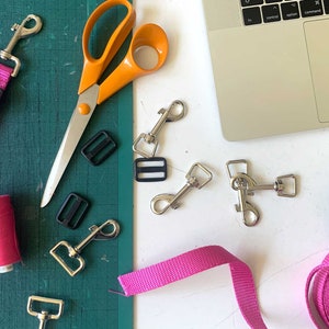 May include: A flat lay of supplies for making dog leashes, including pink and green webbing, silver clasps, black plastic buckles, orange scissors, pink thread, and a ruler.