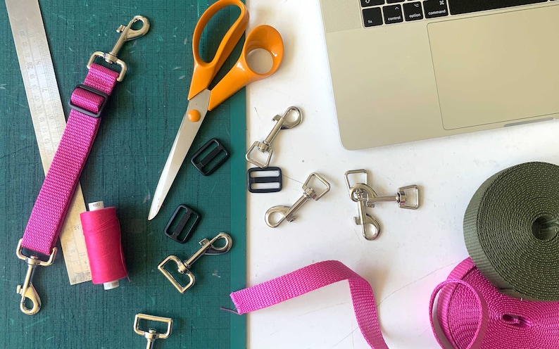 May include: A flat lay of supplies for making dog leashes, including pink and green webbing, silver clasps, black plastic buckles, orange scissors, pink thread, and a ruler.