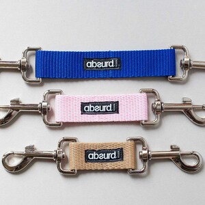 May include: Three dog leash extenders with metal clips and a fabric strap. The straps are blue, pink, and beige. Each strap has the word "abeurd!" printed on it.