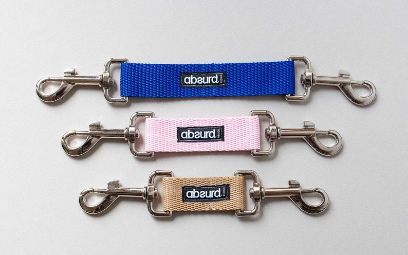 May include: Three dog leash extenders with metal clips and a fabric strap. The straps are blue, pink, and beige. Each strap has the word "abeurd!" printed on it.