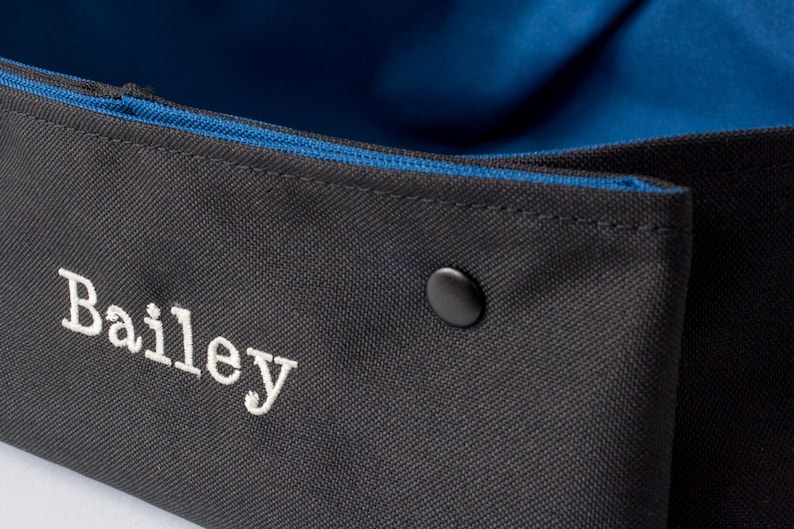 K&ouml;nnte beinhalten: Ein schwarzer Canvas-Aufbewahrungskorb mit blauer Innenausstattung und einem wei&szlig;en gestickten Namen "Bailey".