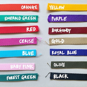 May include: A collection of 14 different colored webbing straps with the color name written in white text below each strap. The colors are orange, yellow, emerald green, purple, red, burgundy, cerise, gold, blue, royal blue, baby pink, olive, forest green, and black.