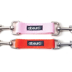 May include: Two dog leash attachments with silver clips. The top attachment is pink with the word "absurd" printed on it. The bottom attachment is orange with the word "absurd" printed on it.