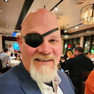 May include: A man wearing a black eye patch over his left eye, a white shirt, and a blue blazer. He has a white beard and mustache. The background shows a restaurant setting with other people seated at tables.