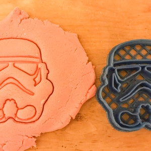 May include: A blue plastic cookie cutter shaped like a Star Wars Stormtrooper helmet. The cutter is on a wooden surface next to a pink cookie dough.