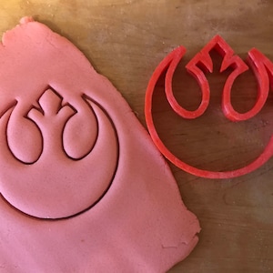 Może przedstawiać: Różowe ciasto na ciasteczka z czerwonym plastikowym wykrawaczem w kształcie symbolu Rebelii z Gwiezdnych Wojen. Wykrawacz leży na cieście.