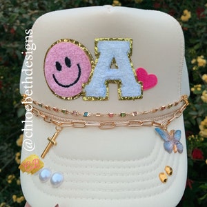 May include: A white trucker hat with a pink smiley face patch, a white felt letter "A" patch with gold glitter, a pink heart patch, a gold chain with a cross charm, and a blue butterfly charm.