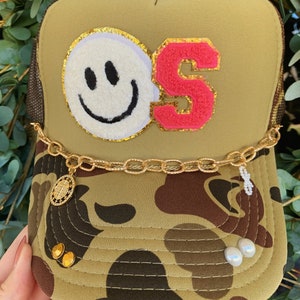 May include: A green camouflage trucker hat with a gold chain and a smiley face and the letter 'S' embroidered on the front.