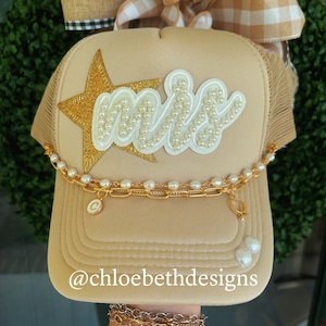 May include: A beige trucker hat with a gold star and the word "Mrs." in white with pearl accents. The hat has a gold chain with a charm attached.