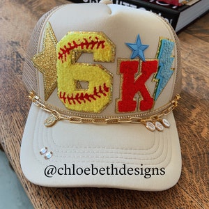 May include: A beige trucker hat with a gold star, a yellow softball with red stitching, a red letter "K", a blue star, and a blue lightning bolt. The hat has a gold chain with charms that spell "B.B.T." and the text "@chloebethdesigns" is on the brim.