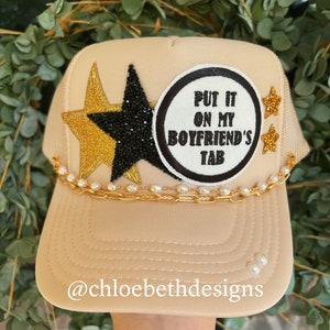 May include: A beige trucker hat with a gold chain, pearls, and a black and gold star. The hat also has a white patch with black text that reads "Put it on my boyfriend's tab".