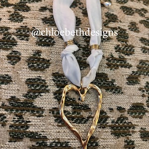 May include: A gold-colored heart pendant necklace with a textured surface. The pendant hangs from a white fabric ribbon, secured with gold rings. The text "@chloebethdesigns" is visible.