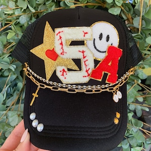 May include: Black trucker hat with a white and red embroidered "5A" patch, a gold star with a red heart, and a smiley face patch. The hat has a gold chain with a cross charm and a gold charm with the letter "L" on it.
