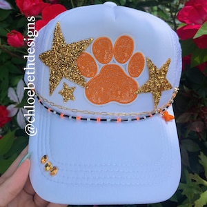 May include: White trucker hat with an orange paw print and gold glitter stars. The hat has a black and white beaded chain with a small orange charm.