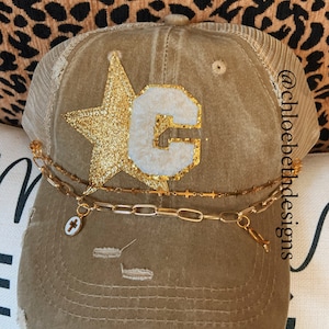 May include: A brown distressed trucker hat with a gold glitter star and a white felt letter "C" on the front. The hat has a gold chain with a cross charm attached to it. The hat is on a leopard print background. @chloebethdesigns