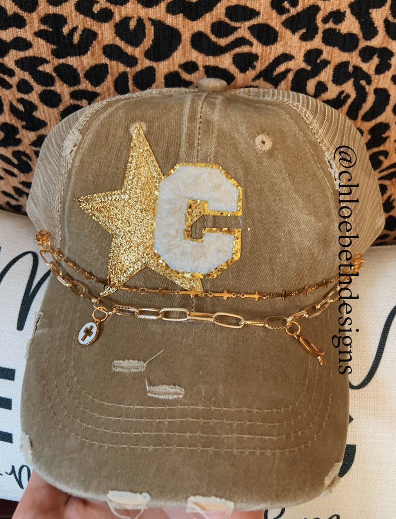 May include: A brown distressed trucker hat with a gold glitter star and a white felt letter "C" on the front. The hat has a gold chain with a cross charm attached to it. The hat is on a leopard print background. @chloebethdesigns