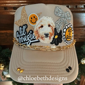May include: A beige trucker hat with a variety of patches, including a dog, a smiley face, a star, a bow, and a flower. The hat also has a gold chain and the text "a lil' bougie" on it. The hat is sitting on a table with a green plant in the background. The text "@chloebethdesigns" is on the bottom of the hat.