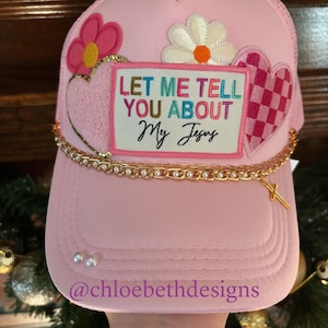 May include: Pink trucker hat with a gold chain, pearls, and patches that say "Let Me Tell You About My Jesus" and feature hearts, flowers, and a checkered pattern. The hat also has a small gold cross charm.