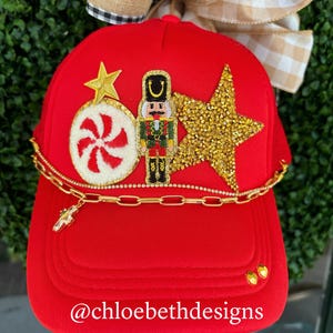 May include: Red trucker hat with a gold chain, a candy cane, a star, and a nutcracker design. The hat has the text "@chloebethdesigns" on the brim.