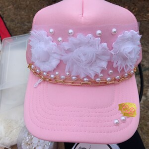 May include: A pink trucker hat with white fabric flowers and pearl accents. A gold chain with pearls is attached to the hat. The hat has a gold sticker with the letters "EB" on it.