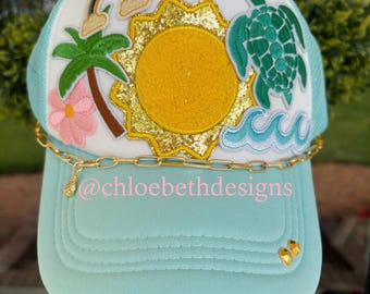 May include: A light blue trucker hat with a white front panel featuring a sun, rainbow, palm tree, flower, and sea turtle design. The hat has a gold chain and the text "@chloebethdesigns".