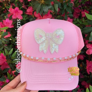 May include: Pink trucker hat with a white sequined bow and a gold chain with white pearls. The hat has the text "@chloebethdesigns" on the side.