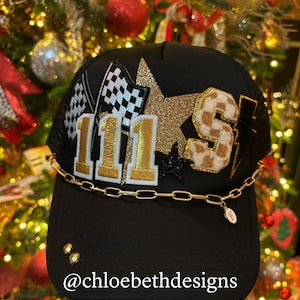 May include: Black trucker hat with a gold chain, gold glitter star, and patches that say "111" and "S". The hat has a checkered flag patch and a lightning bolt patch. The text "@chloebethdesigns" is on the bottom of the hat.