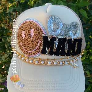 May include: Cream-colored trucker hat with a tan mesh back. The hat features a leopard print smiley face with pink lightning bolts, the word "MAMA" in black, and a silver sequined bow. A gold chain with pearls and a charm adorns the brim.