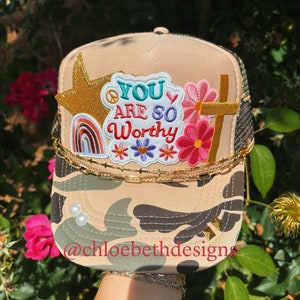 May include: A beige camouflage trucker hat with a gold chain and various colorful patches. The patches include a rainbow, a peace sign, a star, flowers, and a cross. The text on the hat reads "You are so worthy".