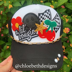 May include: Black and white trucker hat with a gold chain and decorative patches. Patches include a red flower, a smiley face, cherries, a star, and checkered flags. The text "a lil' bougie" is also featured on the hat. The brim has three decorative pearls.