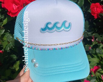 May include: A light blue trucker hat with a white mesh back. The hat has a white front with a blue wave design and a gold chain with colorful beads around the brim. The hat has two white pearl embellishments on the front.