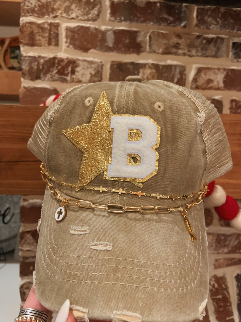 May include: A distressed brown baseball cap with a gold star and a white letter "B" on the front. The cap has a gold chain with a cross charm attached.