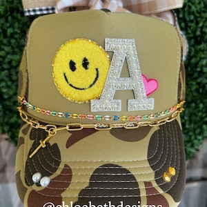 May include: A camouflage trucker hat with a yellow smiley face, a rhinestone-encrusted letter "A", and a pink heart. The hat has a gold chain with a cross charm and a multi-colored chain around the brim. The hat is held in a hand with the text "@chloebethdesigns" visible below the hat.