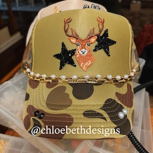 Pu&ograve; includere: Un cappello da camionista mimetico con una catena dorata e un bordo di perle. Il cappello presenta un cervo ricamato con due stelle nere come ornamento. Il testo '@chloebethdesigns' &egrave; visibile sul cappello.