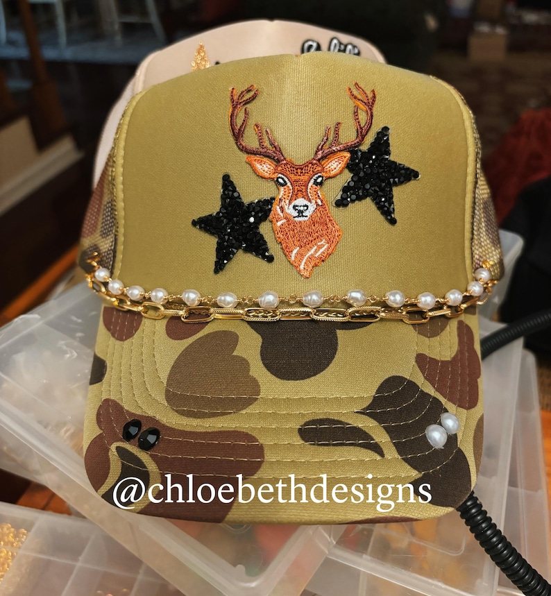 Pu&ograve; includere: Un cappello da camionista mimetico con una catena dorata e un bordo di perle. Il cappello presenta un cervo ricamato con due stelle nere come ornamento. Il testo '@chloebethdesigns' &egrave; visibile sul cappello.