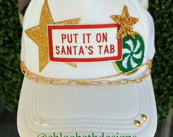 May include: White trucker hat with a gold star, a gold star with a red and white "Put it on Santa's Tab" patch, a green and white peppermint candy patch, and a gold chain. The hat has two pearl-like embellishments on the brim.