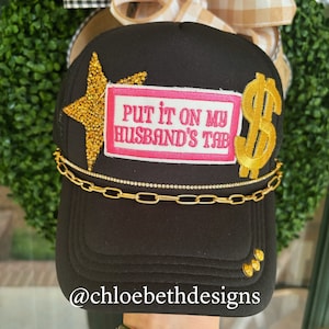 May include: Black trucker hat with a gold star, a pink patch that says "Put it on my husband's tab" and a gold dollar sign. The hat has a gold chain around the brim. The hat is on a table with a hand wearing gold bracelets.