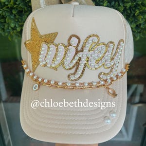 May include: Cream-colored trucker hat with the word "wifey" in pink and gold glitter, adorned with pearls and a gold star. A gold chain with a charm and a ring pendant hangs across the brim. The text "@chloebethdesigns" is visible.