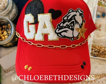 May include: Red trucker hat with a gold chain, black and white bulldog patch, black heart, and white letters that say "GA".