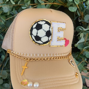May include: A tan trucker hat with a gold chain and a charm. The hat has a white and black soccer ball patch and a white letter E with gold trim. The letter E has a pink heart on it.