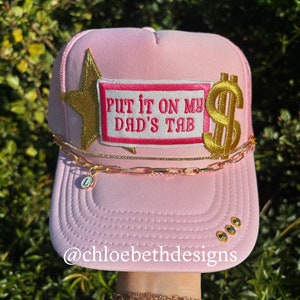 May include: Pink trucker hat with a gold star, a gold dollar sign, and a pink and white patch that says "Put it on my dad's tab".