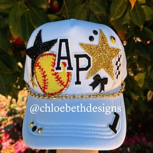 May include: White baseball cap with a gold star, black star, softball, and a gold chain. The cap has the letters "AP" in black and white on the front and the number "1" on the side. The text "@chloebethdesigns" is on the front of the cap.