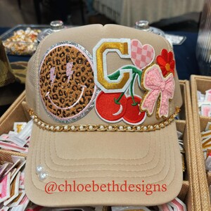 May include: A beige trucker hat with a variety of colorful patches, including a smiley face, a letter G, a cherry, a heart, and a bow. The hat has a gold chain and pearls around the brim. The text "@chloebethdesigns" is printed on the front of the hat.
