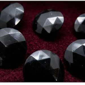 Può includere: Una collezione di bottoni neri rotondi caratterizzati da una superficie geometrica sfaccettata. Ogni bottone presenta una finitura lucida e riflettente con molteplici sfaccettature triangolari che creano un aspetto angolare sulla forma circolare.