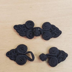 Puede incluir: Tres cierres de rana negros, también conocidos como nudos chinos, se muestran sobre una superficie de madera clara. Los cierres están hechos de cordón tejido y presentan un diseño en bucle. Estos cierres decorativos se utilizan a menudo en la ropa.
