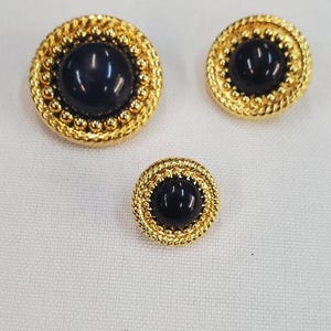 Puede incluir: Tres botones dorados con un centro negro de forma ovalada. Los botones tienen un borde texturizado en forma de cuerda. Los botones son de tres tamaños diferentes, con el botón más grande a la izquierda y el más pequeño en la parte inferior.