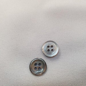 Puede incluir: Dos botones redondos e iridiscentes con cuatro agujeros cada uno, sobre una tela beige claro. Los botones tienen un brillo nacarado sutil, y la tela tiene una textura finamente tejida. Adecuado para costura o manualidades.