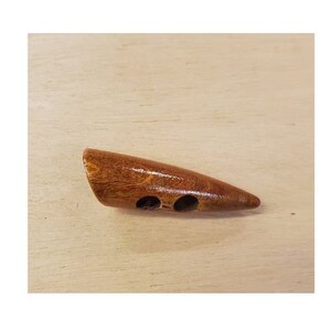 Puede incluir: Un botón de madera con acabado marrón brillante. El botón es alargado con un extremo redondeado y dos agujeros para coser. El botón está sobre una superficie de madera clara.